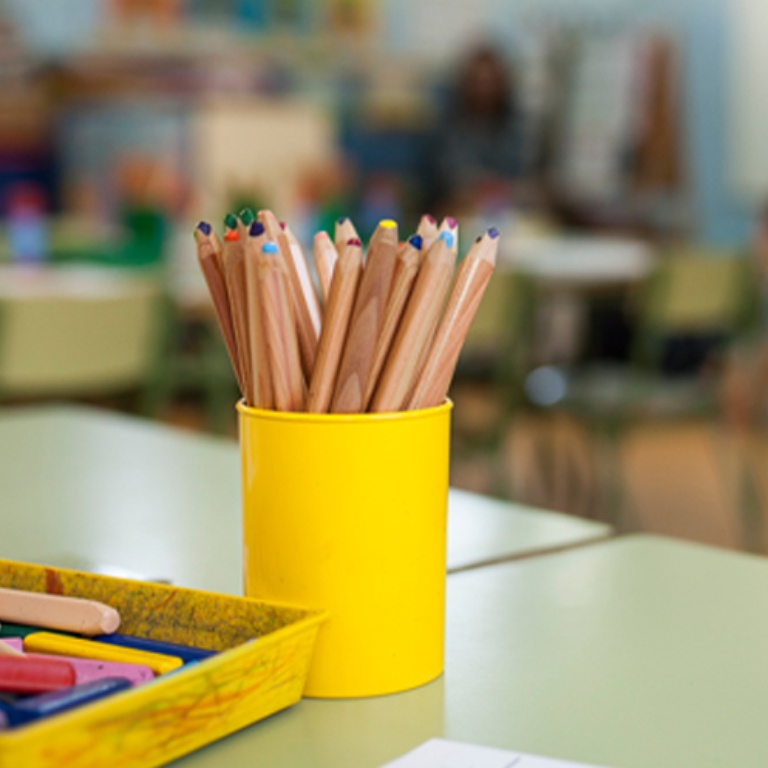 A cup of colored pencils and a scribbled-on tray of crayons on a desk in the front of a classroom