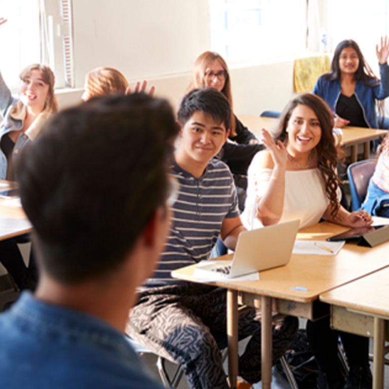 Male high school teacher standing at front of class, teaching while students raise their hands.