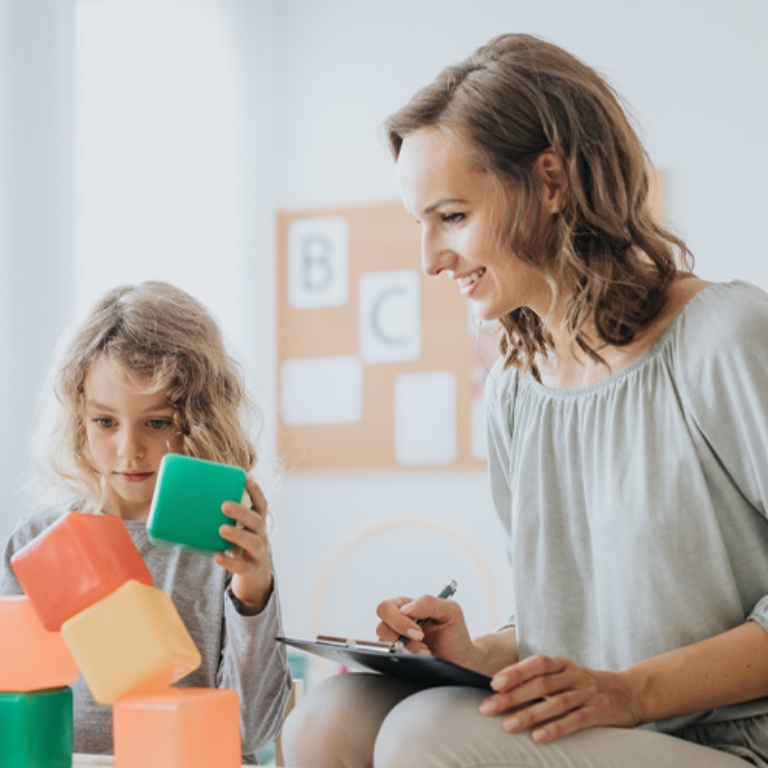 Young school psychologist holds a clipboard and watches as young student works with large foam blocks in a session.